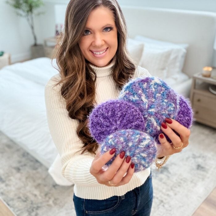 A woman stands in a bedroom, smiling at the camera as she holds several round, knitted purple and multicolored Crochet Scrubbies in her hands.
