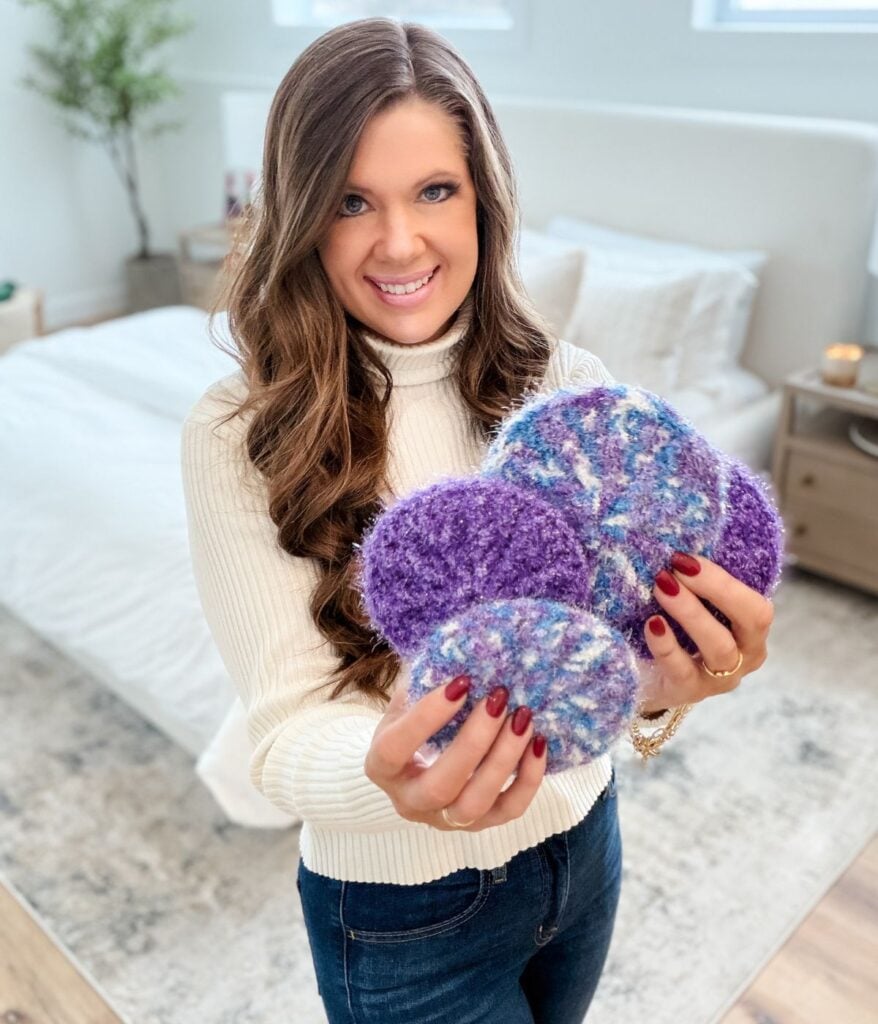 A woman stands in a bedroom, smiling at the camera as she holds several round, knitted purple and multicolored Crochet Scrubbies in her hands.