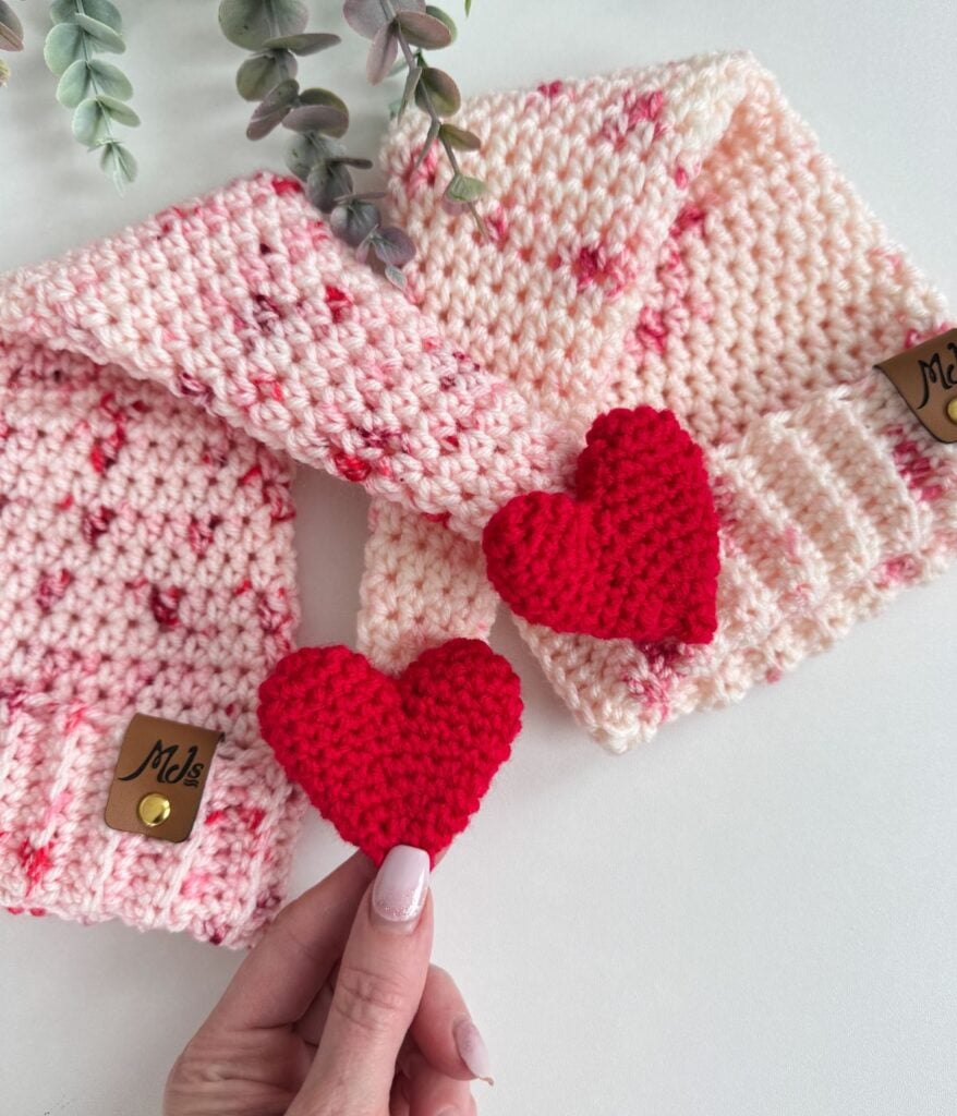 Two pink knitted hats with speckled patterns and brown tags are displayed on a white surface; a hand holds a red crocheted heart beside another heart placed on the hats, creating a charming Valentine’s Crochet scene.