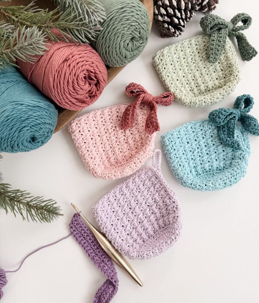 Four crocheted square cloths in pastel colors, perfect as mug cozies, with bows, yarn balls, a crochet hook, pinecones, and evergreen branches arranged on a white surface.