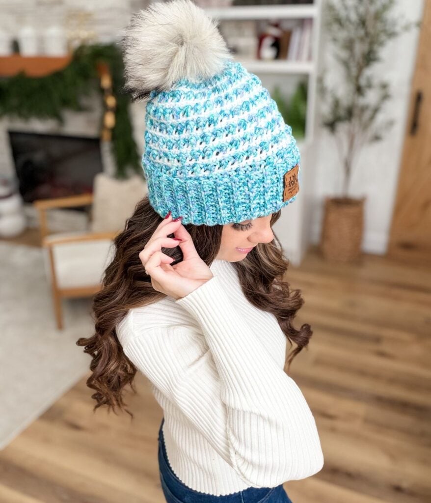 A woman with long brown hair wears a white ribbed sweater and a blue Winterberry Beanie, standing in a cozy, well-lit room with wood floors and plants—perfect inspiration for your next Free Crochet Pattern project.
