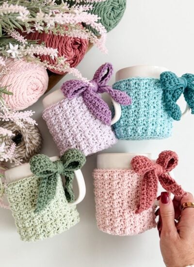 Four white mugs with pastel-colored mug cozies, each tied with a matching bow, are arranged near yarn balls and flowers on a white surface. A hand holds the pink mug cozy.