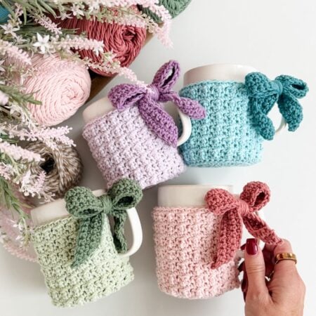 Four white mugs with pastel-colored mug cozies, each tied with a matching bow, are arranged near yarn balls and flowers on a white surface. A hand holds the pink mug cozy.