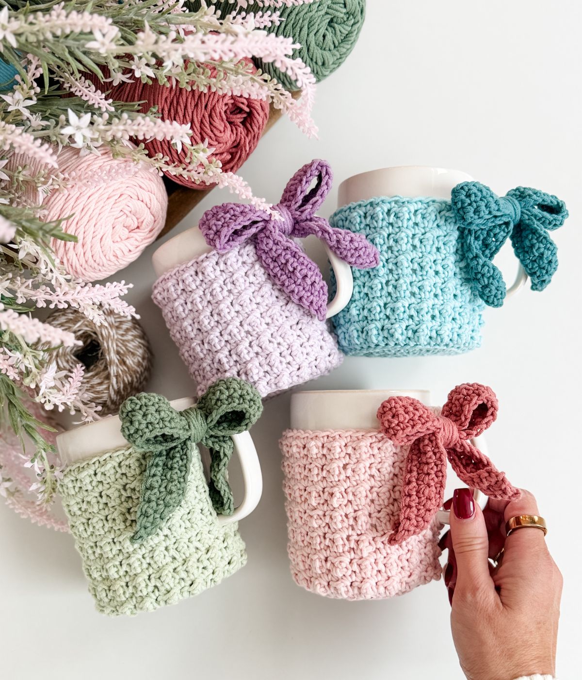 Four white mugs with pastel-colored mug cozies, each tied with a matching bow, are arranged near yarn balls and flowers on a white surface. A hand holds the pink mug cozy.