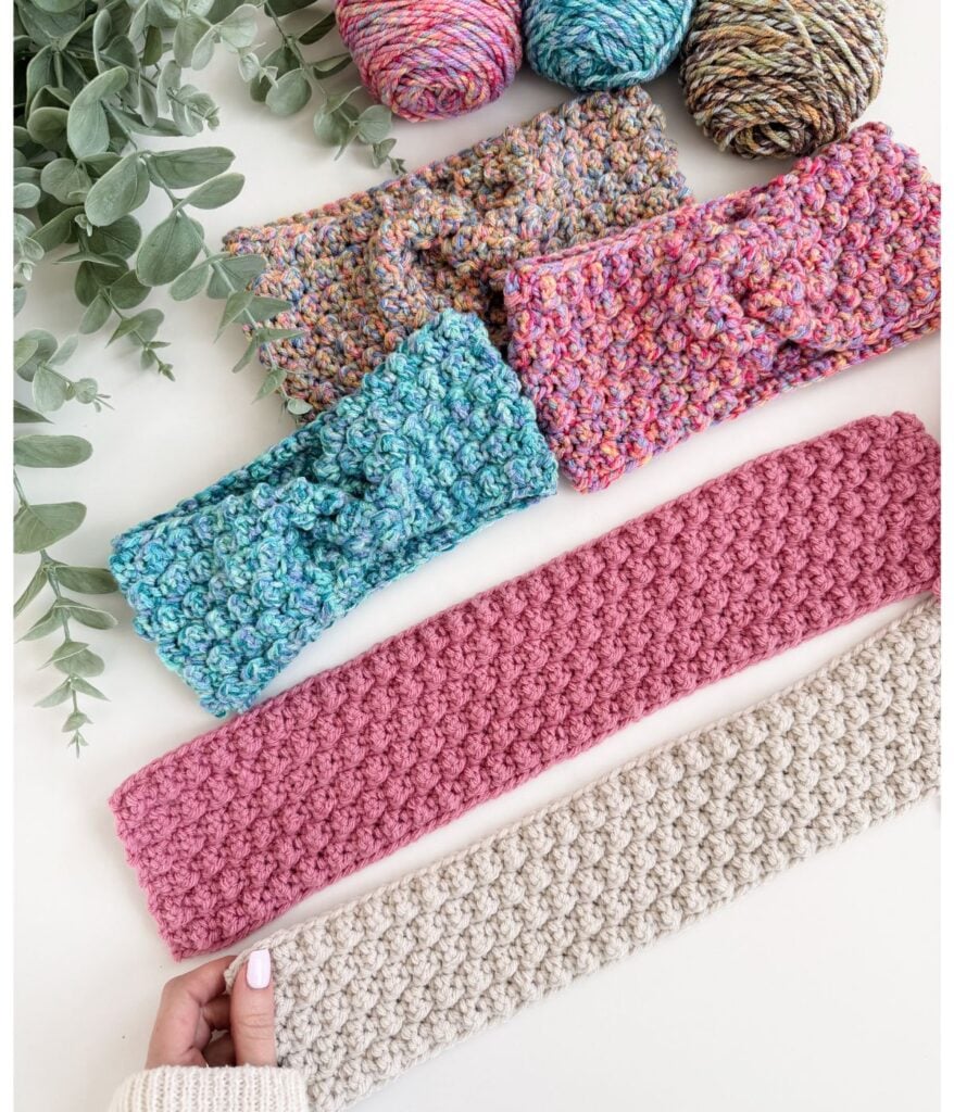 Four crocheted ear warmer headbands in various colors are displayed on a white surface next to green foliage and balls of multicolored yarn, with a hand holding one headband.