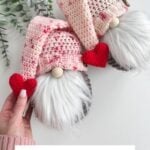 Two crocheted Valentines Gnomes with pink hats, white beards, and red hearts are held in a hand, framed by eucalyptus leaves. Text reads, "Rustic Farmhouse Valentines Gnome.