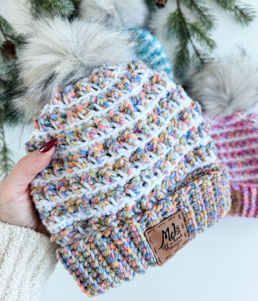 A hand holds a multicolored Winterberry Beanie crochet beanie with a faux fur pom-pom and a leather tag that reads "MJs Off The Hook Designs"; other similar beanies are in the background. Free crochet pattern available.