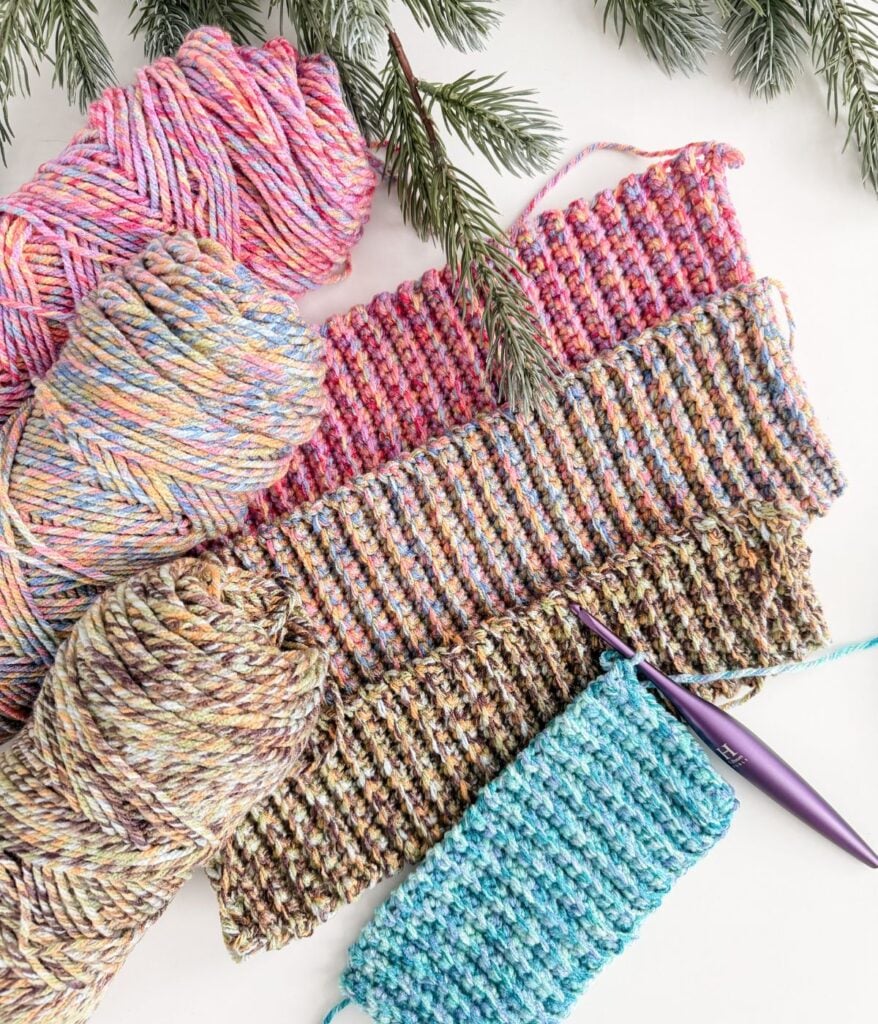 Four crocheted swatches in varying multicolored yarns, a purple crochet hook, and yarn balls are arranged on a white surface with pine branches above—perfect inspiration for your next Crochet Beanie or Winterberry Beanie project.