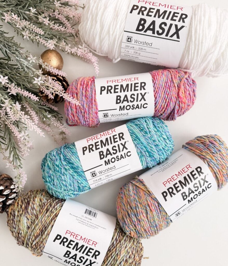 Four skeins of Premier Basix Mosaic yarn in various colors and one skein of white Premier Basix yarn are arranged beside pine branches and pine cones—a cozy setup perfect for creating the Winterberry Beanie Free Crochet Pattern.
