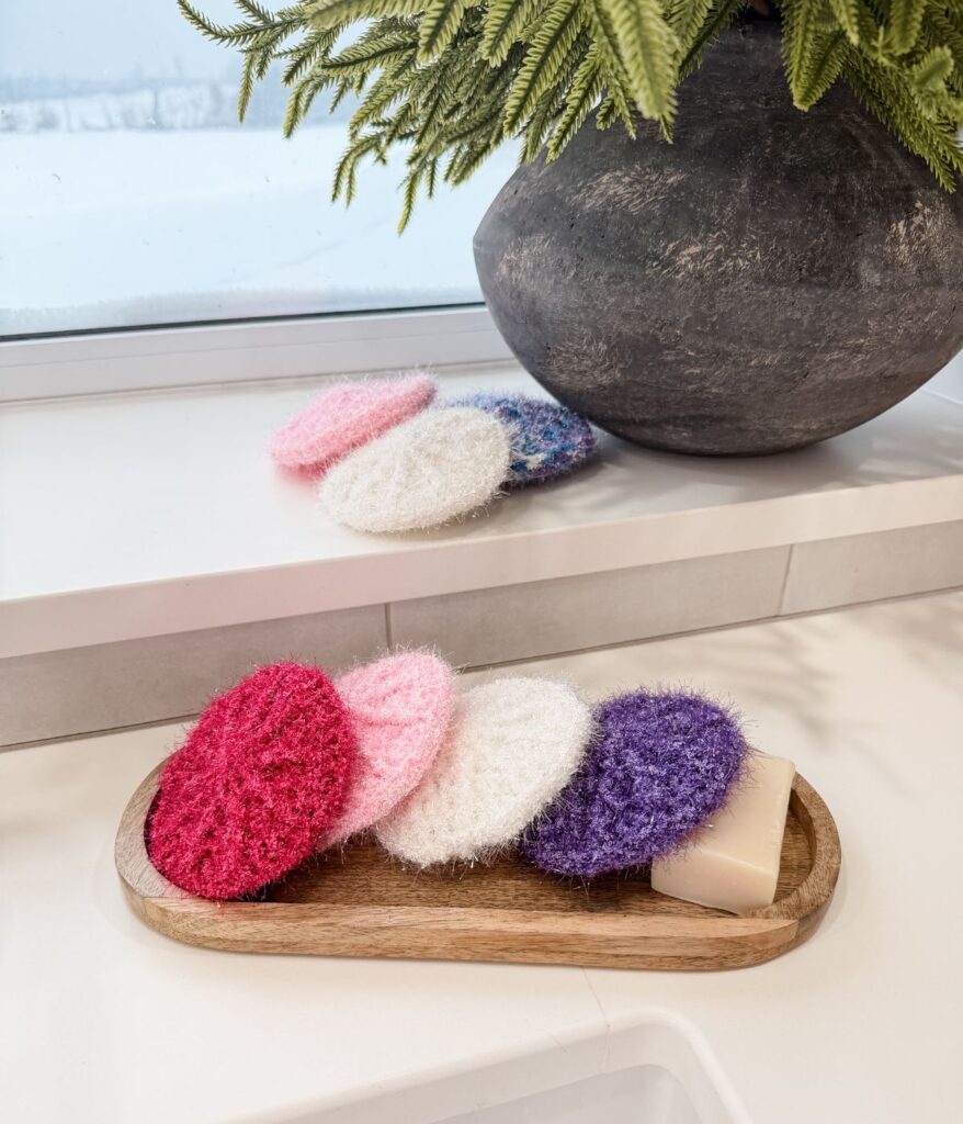 A wooden tray holding a bar of soap and several round, colorful crochet scrubbies sits on a white countertop near a window and a large vase with green foliage.