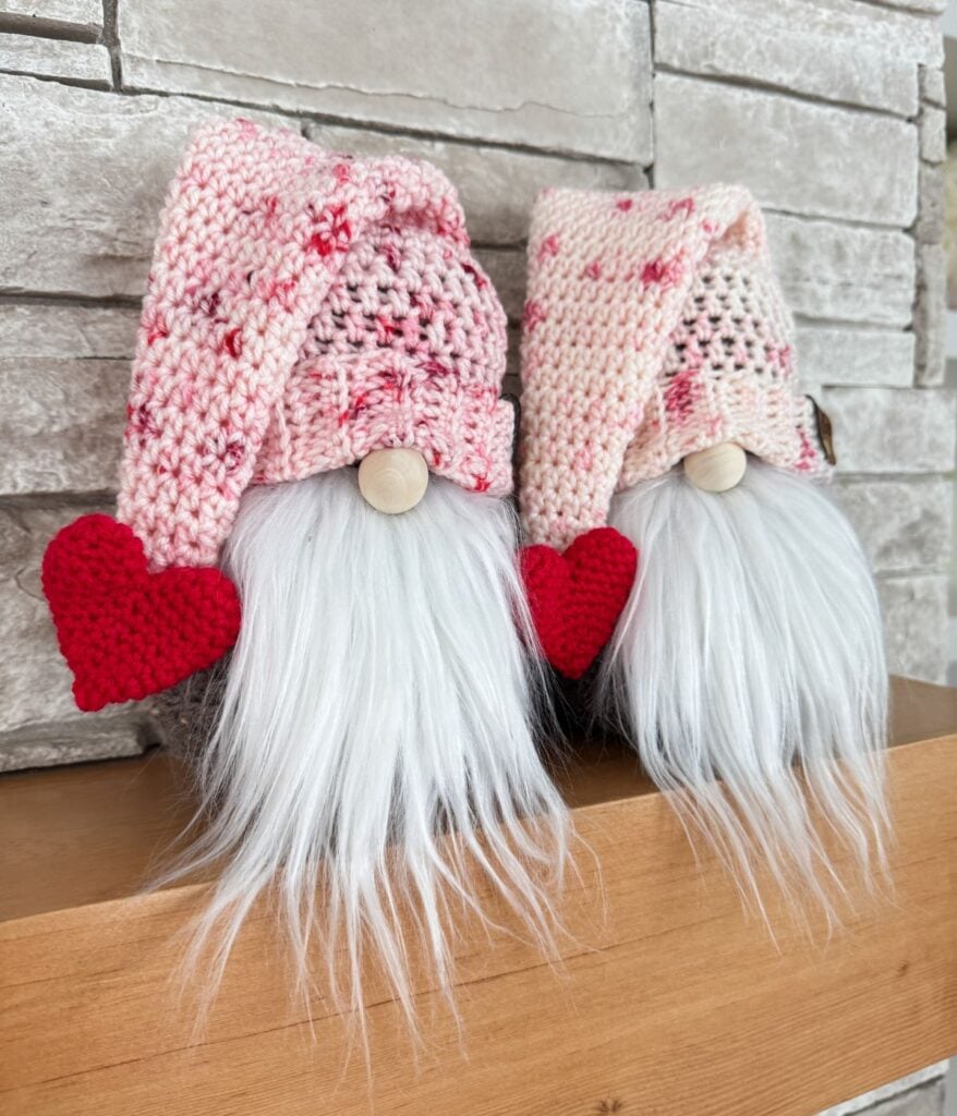 Two gnome figurines with white beards, pink knit hats, and red heart decorations sit side by side on a wooden surface in front of a stone wall—a charming Valentine’s Gnome scene perfect for Valentine’s Crochet inspiration.
