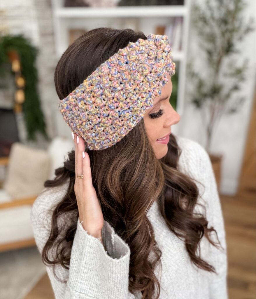 A woman with long brown hair wears a chunky, multicolored crocheted ear warmer headband and a light gray sweater, touching the ear warmer with one hand indoors.