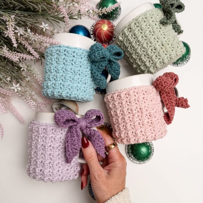 Four mugs with pastel Mug Cozy wraps and bows are arranged among Christmas ornaments and plants; a hand holds the lilac Mug Cozy in the foreground.
