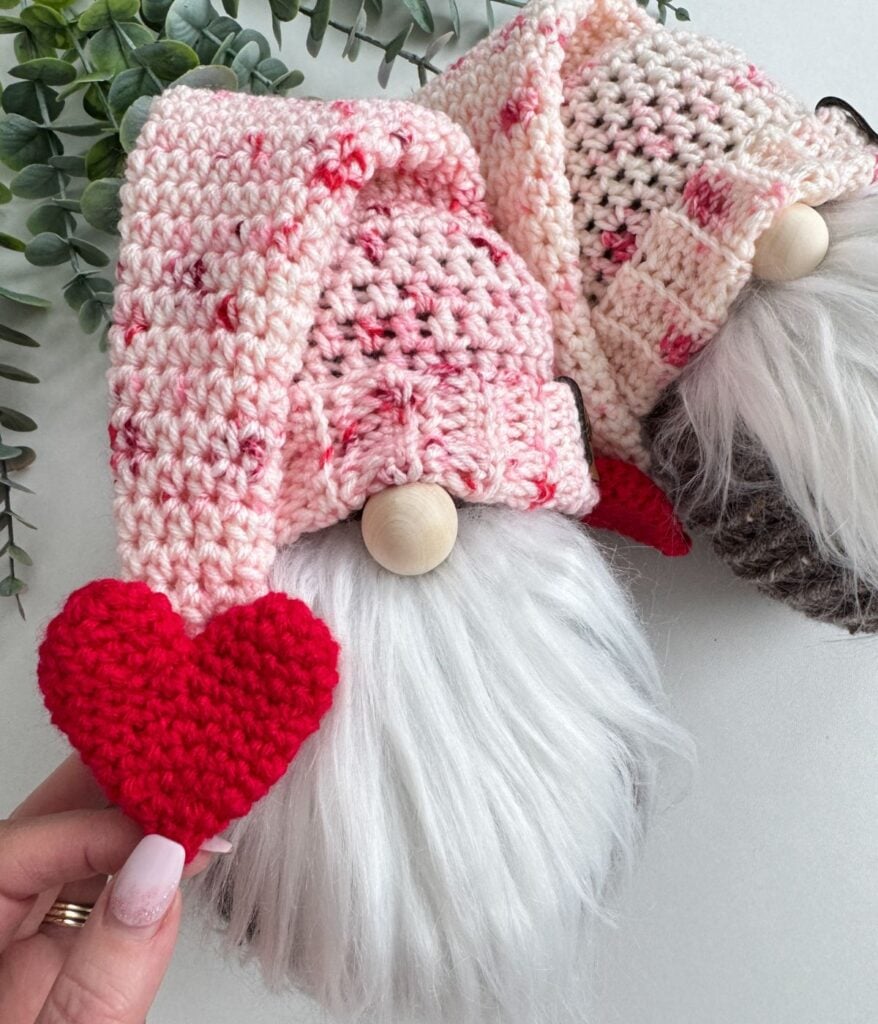 Two crocheted Valentine’s Gnome figures with white beards, pink hats, and wooden noses; one features a red crocheted heart on its hat. Greenery decor is in the background—a charming Valentine’s Crochet accent.
