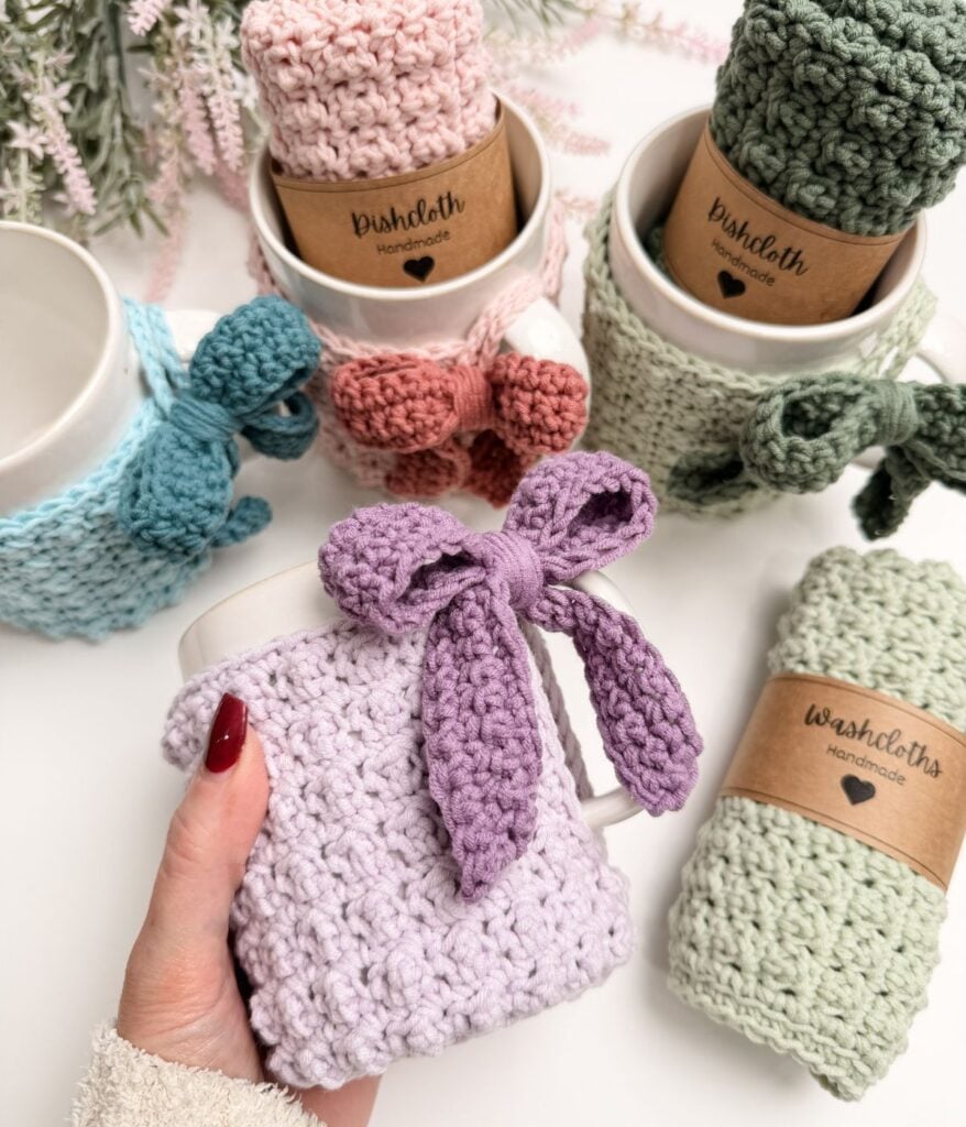 Hand holding a white mug wrapped in a light purple crocheted mug cozy with a bow, surrounded by other mugs and rolled crocheted dishcloths in pastel colors.