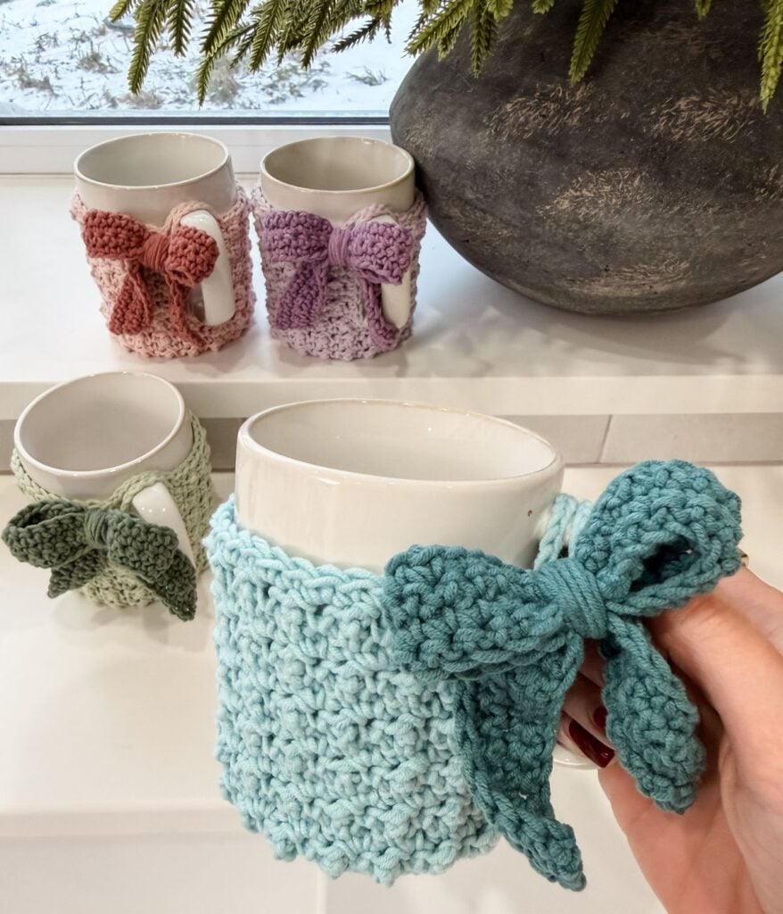 Four white mugs, each wrapped in a pastel Mug Cozy with a large bow, create a charming display. In the foreground, a hand holds the mug adorned with a turquoise Mug Cozy for an extra touch of warmth and style.