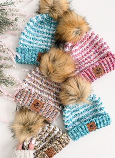Five Winterberry Beanies with faux fur pom-poms and multicolored patterns are arranged on a white surface next to light pink artificial foliage, perfect for trying a new crochet pattern this season.