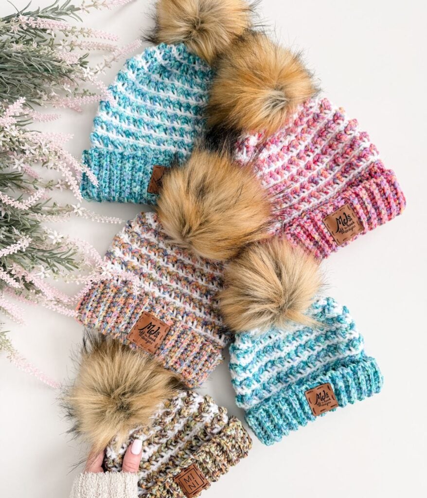 Five Winterberry Beanies with faux fur pom-poms and multicolored patterns are arranged on a white surface next to light pink artificial foliage, perfect for trying a new crochet pattern this season.