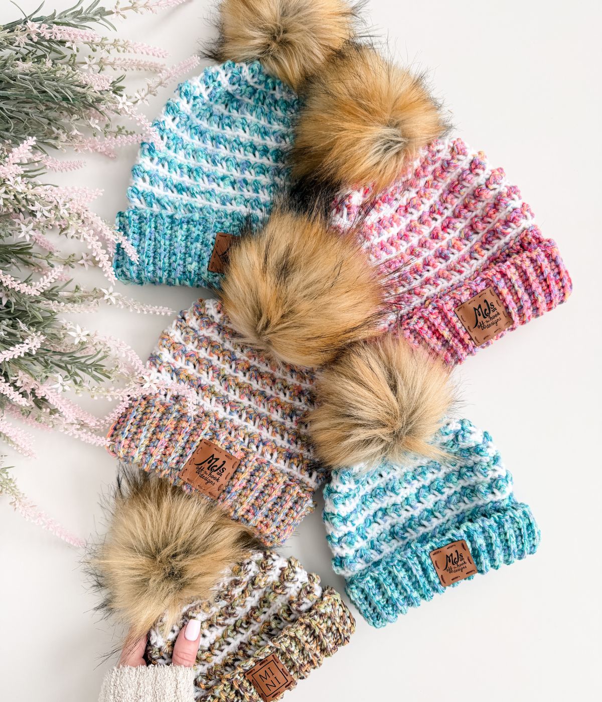 Five Winterberry Beanies with faux fur pom-poms and multicolored patterns are arranged on a white surface next to light pink artificial foliage, perfect for trying a new crochet pattern this season.