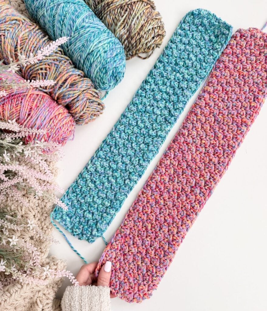 Two crocheted scarves, one blue and one purple-pink, are displayed with matching yarn skeins and an ear warmer on a white surface next to a bunch of artificial flowers.