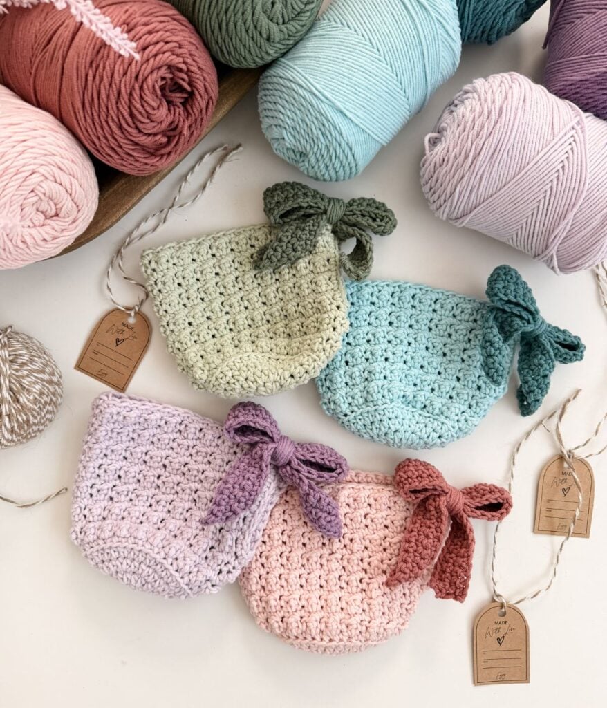 Four crocheted mini pouches in pastel colors, each with a matching bow, are displayed near rolls of yarn and handmade tags on a white surface—perfect for gifting or storing small items like a mug cozy.