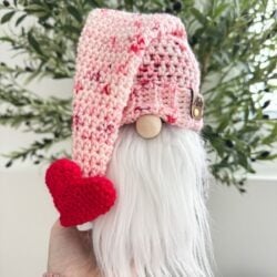 A hand holds a small Valentine’s Gnome figurine with a white beard, pink crocheted hat, and red crocheted heart, in front of green leafy plants.