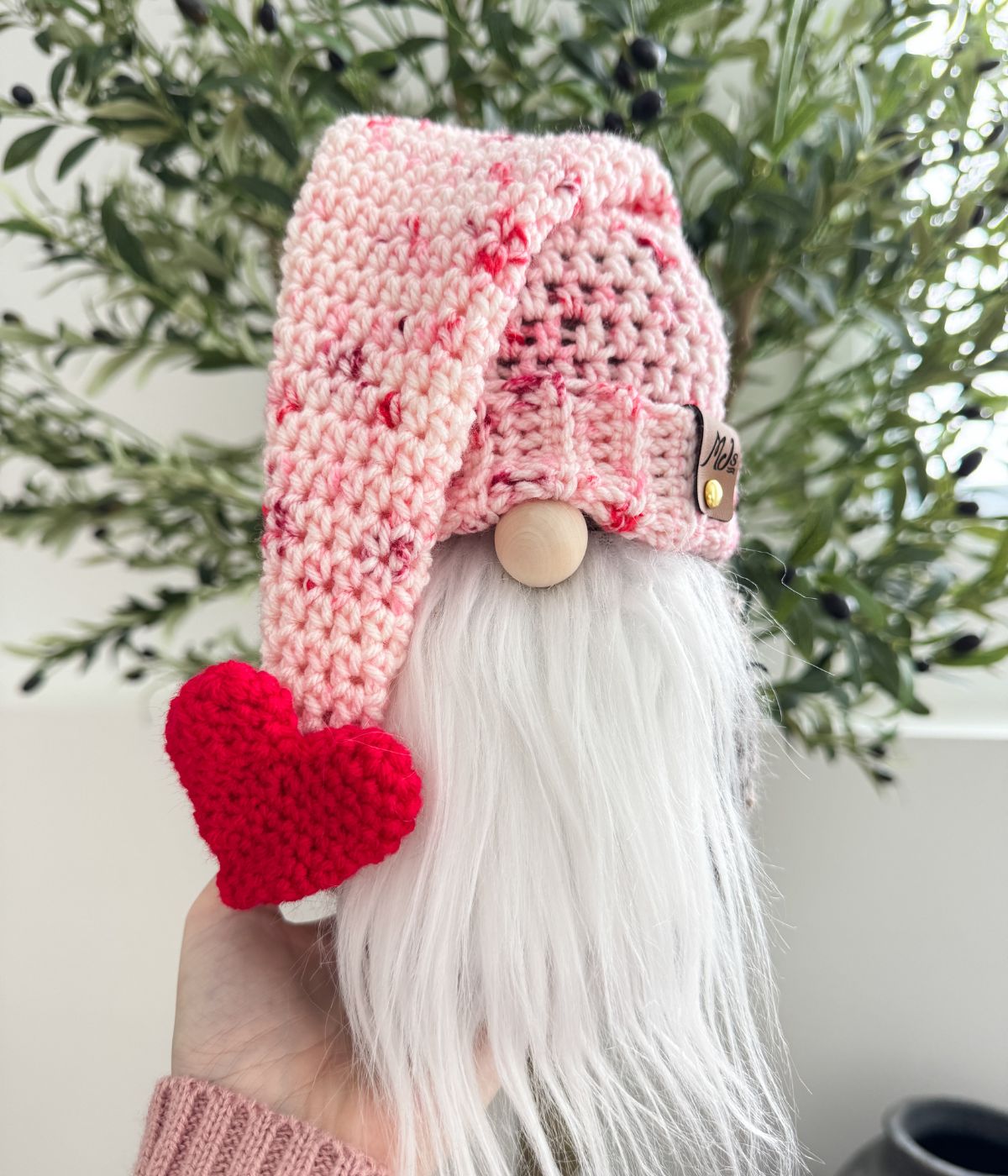 A hand holds a small Valentine’s Gnome figurine with a white beard, pink crocheted hat, and red crocheted heart, in front of green leafy plants.