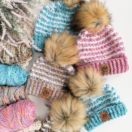 Four knitted hats with faux fur pom-poms are arranged among skeins of colorful yarn and a sprig of faux greenery on a white surface, showcasing the cozy Winterberry Beanie—a free crochet pattern perfect for winter.
