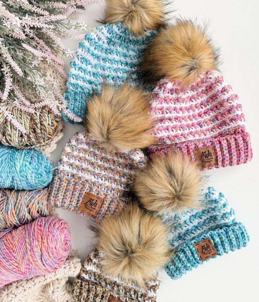 Four knitted hats with faux fur pom-poms are arranged among skeins of colorful yarn and a sprig of faux greenery on a white surface, showcasing the cozy Winterberry Beanie—a free crochet pattern perfect for winter.