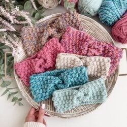 A round tray holds six crocheted ear warmer headbands in various colors and textures, surrounded by yarn balls and greenery. A hand reaches toward a blue ear warmer at the front.