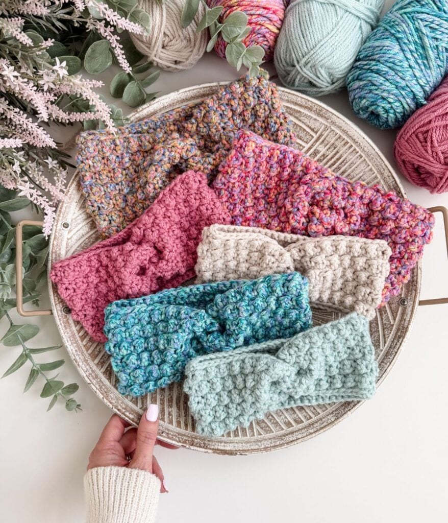 A round tray holds six crocheted ear warmer headbands in various colors and textures, surrounded by yarn balls and greenery. A hand reaches toward a blue ear warmer at the front.