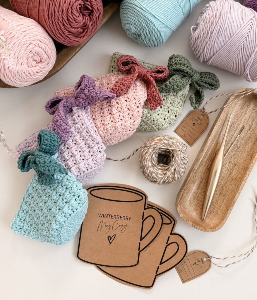 Assorted pastel crocheted pouches with bows, yarn balls, crochet hooks, twine, and two brown Mug Cozy tags labeled "WINTERBERRY My Cozy" arranged on a white surface.