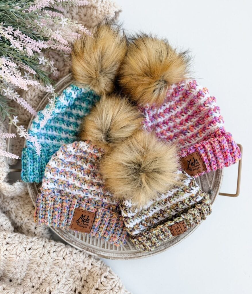 A woven basket holds four colorful crochet beanies, each with a large faux fur pom-pom on top, placed on a white surface near a plant. Find your new favorite Winterberry Beanie made with a free crochet pattern.