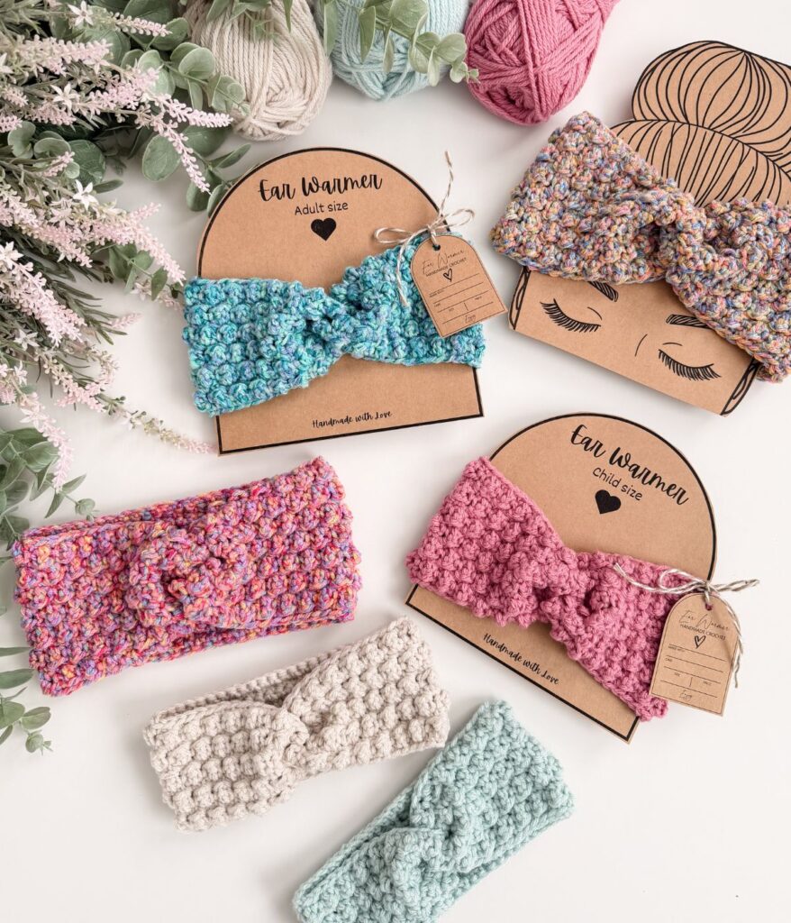 Four stylish ear warmers in pink, blue, and cream are crocheted with care and displayed on cardboard holders, surrounded by decorative yarn balls and faux greenery.