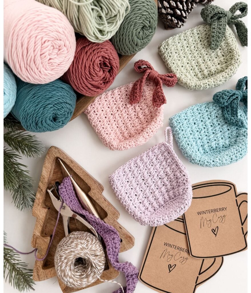 Assorted yarn, Mug Cozy crafting tools, crocheted mug cozies, and “Winterberry Mug Cozy” tags are arranged on a white surface with pine accents.