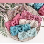 A tray displays five colorful crocheted ear warmers, yarn balls, crochet hooks, and a plant, with text promoting a cozy Ear Warmer pattern perfect for winter crafting.