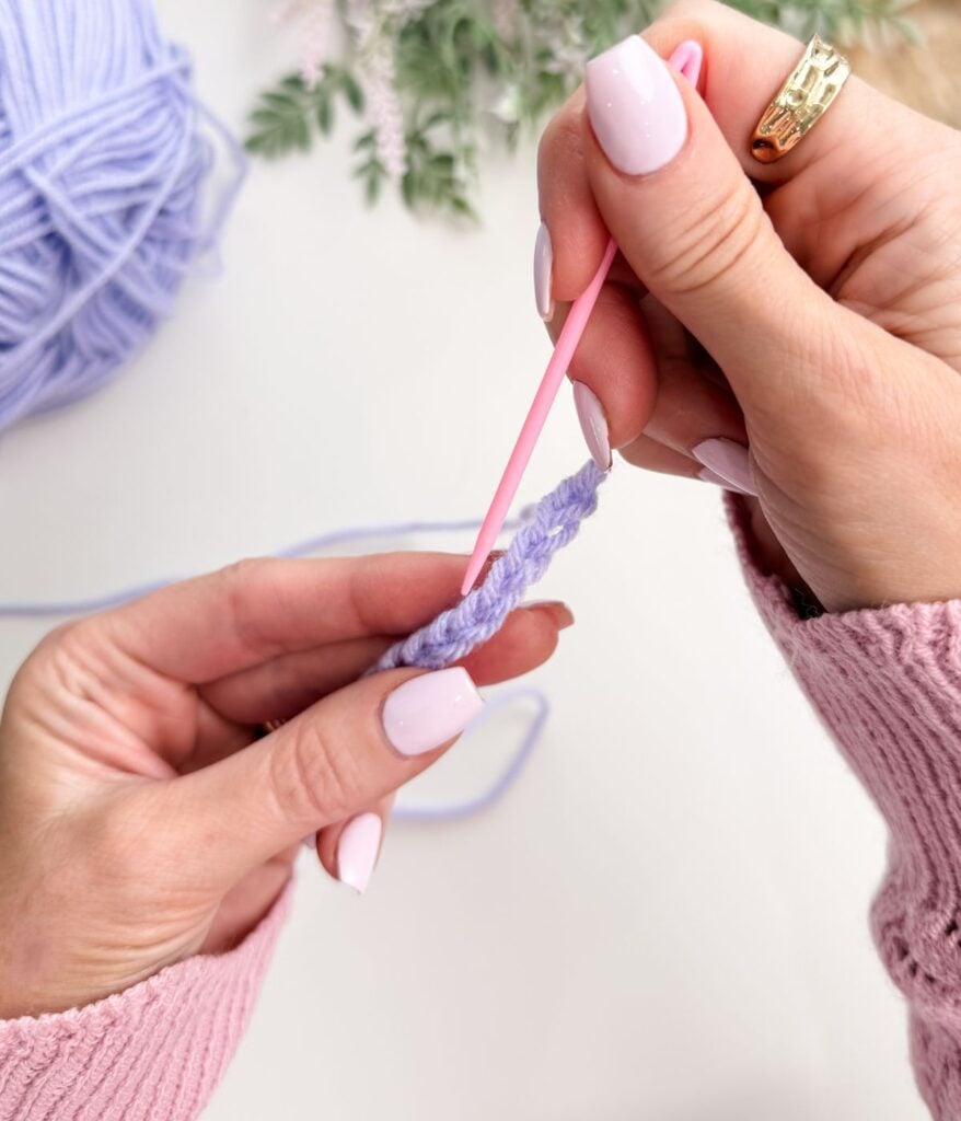 Close-up of hands using a pink plastic yarn needle to sew a row of light purple yarn, with a slip knot and chain stitch starting the work. A ball of yarn and greenery are visible in the background.