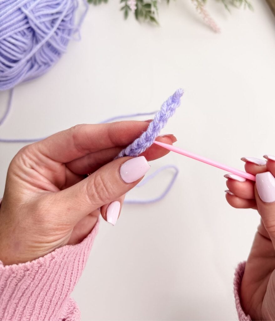 Hands holding a pink yarn needle threading purple yarn, ready to start a slip knot and chain stitch, with a ball of yarn and some flowers in the background.