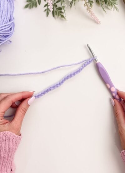 Hands holding a crochet hook and purple yarn, forming a slip knot and working chain stitches, with flowers and a ball of yarn in the background on a white surface.