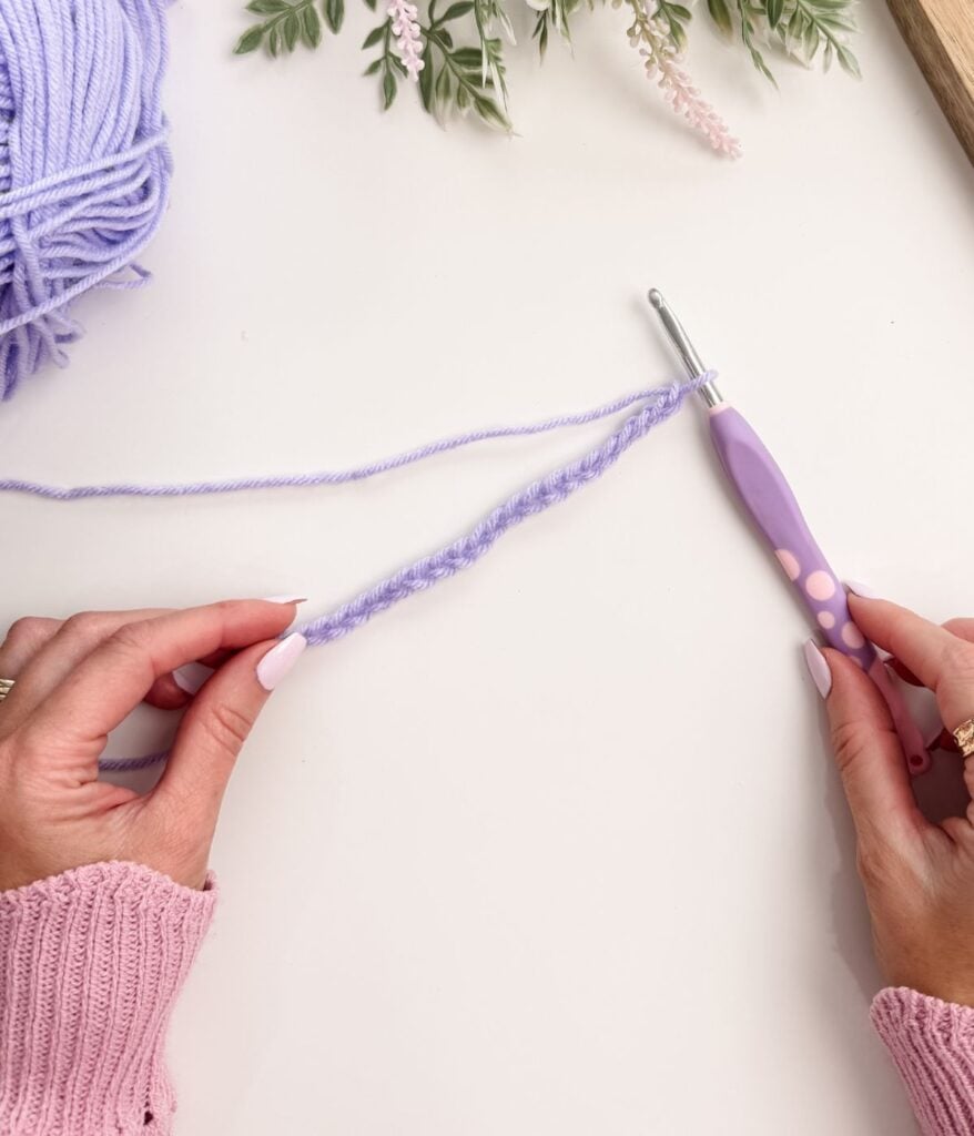 Hands holding a crochet hook and purple yarn, forming a slip knot and working chain stitches, with flowers and a ball of yarn in the background on a white surface.
