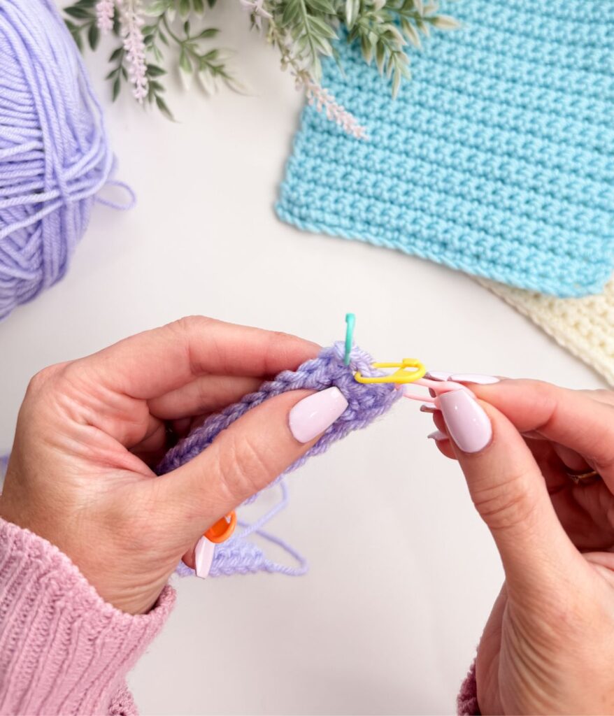 A person crochets with lavender yarn, using stitch markers and practicing single crochet for beginners, while blue and cream crocheted swatches and a ball of yarn are visible nearby.