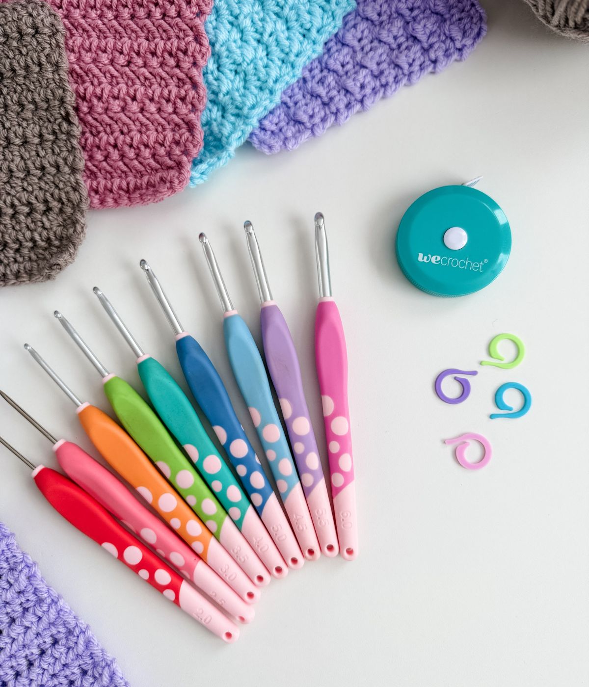 A set of colorful crochet hooks, yarn swatches, a teal tape measure, and three plastic stitch markers on a white surface.