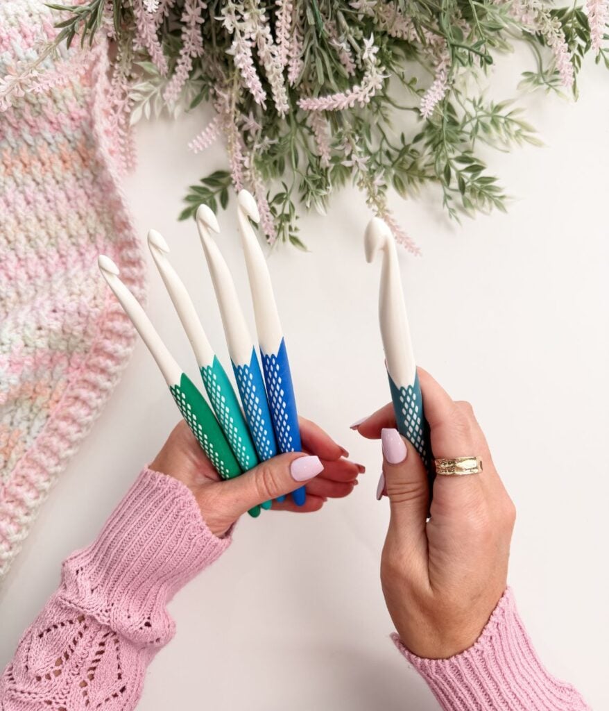 A person wearing a pink sweater holds six large crochet hooks with blue and green handles; a crocheted item and greenery are on the table.