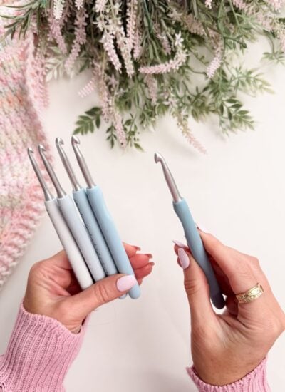 A person holds four crochet hooks in one hand and one in the other, with a knitted fabric and greenery in the background.