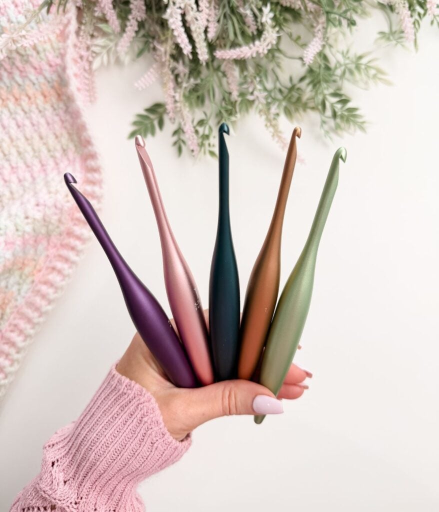 A hand holds five ergonomic crochet hooks of different colors. A crochet piece and greenery are in the background.
