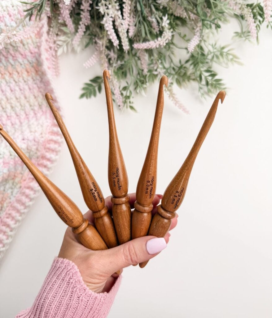 A hand holding five wooden crochet hooks of various sizes, with a leafy plant and a crocheted fabric in the background.