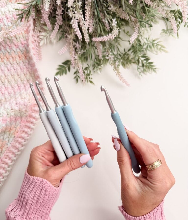 A person holds four crochet hooks in one hand and one in the other, with a knitted fabric and greenery in the background.