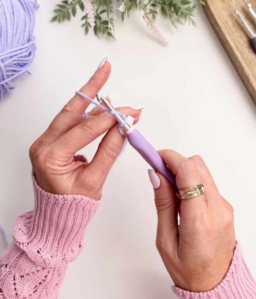 Close-up of hands in a pink sweater crocheting a slip knot and chain stitch with a lavender crochet hook and light purple yarn on a white surface.