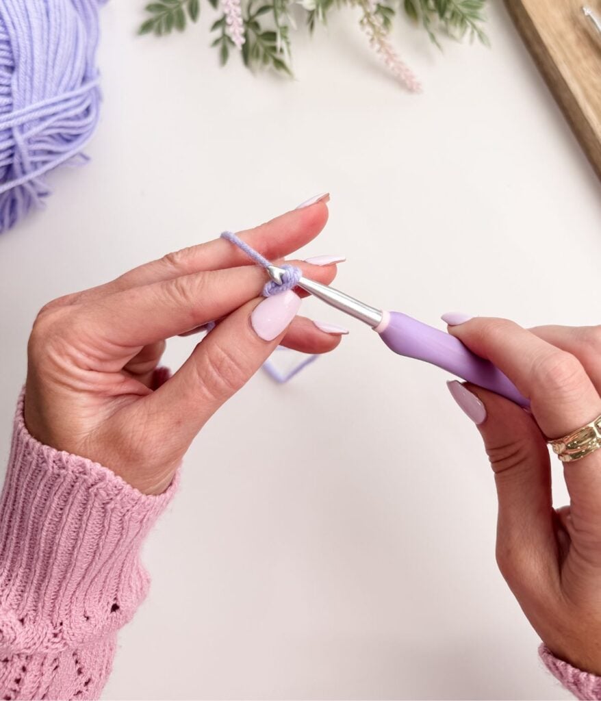 Close-up of hands crocheting with light purple yarn and a crochet hook, starting with a slip knot and chain stitch, with a ball of yarn and greenery visible in the background.
