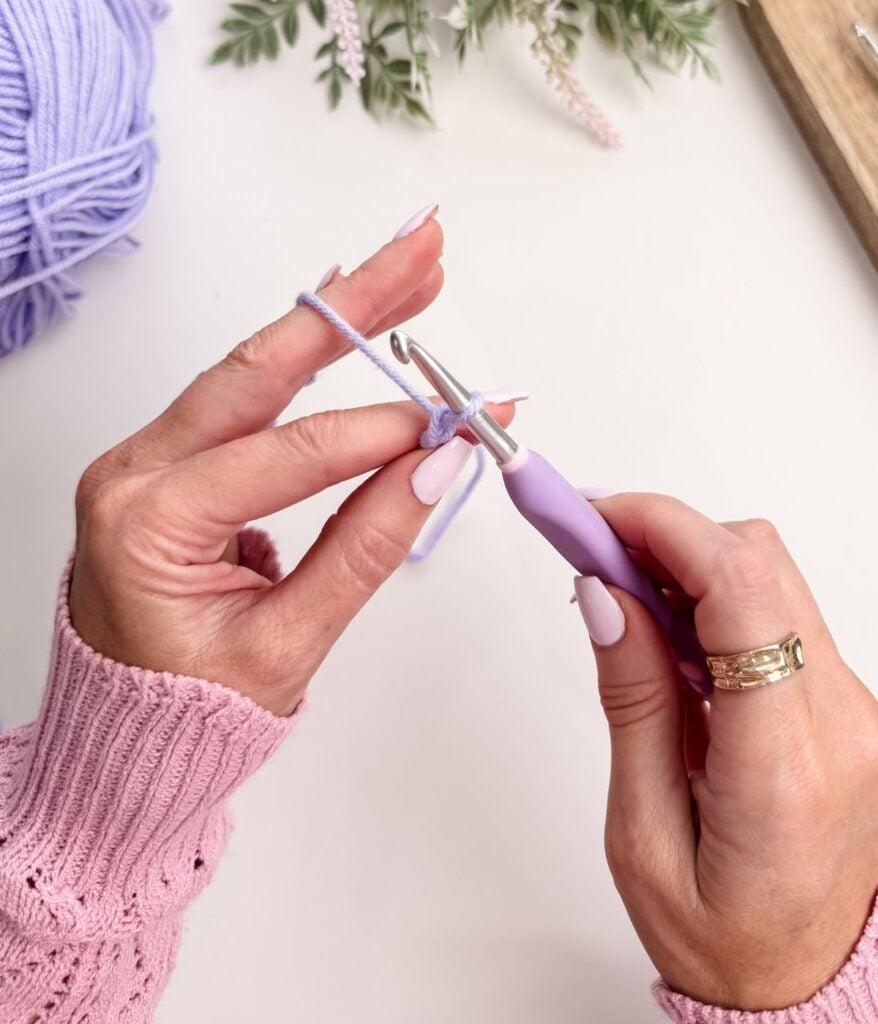 Close-up of hands crocheting a slip knot and chain stitch with lavender yarn and a purple crochet hook, wearing a pink sweater and gold ring, with a yarn ball and flowers in the background.